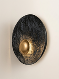 A circular wall sconce with an attractive contrasting design, featuring a black background with a rough stone texture and a smooth brass dome that reflects warm backlighting.