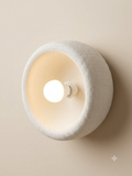 A circular wall sconce with a hollow design and a rough finish resembling plaster or pottery, with a spherical bulb visible in the center.