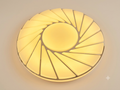 A circular ceiling light unit with a spiral radial design resembling sunlight or a turbine fan, providing warm lighting.