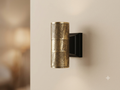 A cylindrical wall light unit with an antique brass finish and embossed patterns that give it a classic character, mounted on a black base.
