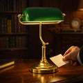 The Banker's Lamp, a classic desk lamp, features a polished brass base and stand and an iconic green glass shade, giving it a vintage feel and providing focused, comfortable reading light.